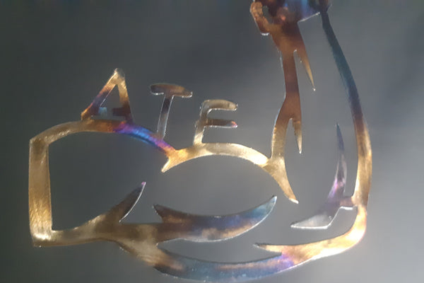 A metal wall art piece featuring the word 'ATF' in stylized cutout letters, with an additional abstract curved shape rising above the letters. The metal has an iridescent gold finish with rainbow color shifts, shown against a dark gray background.