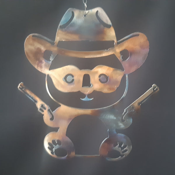A metal wall hanging shaped like a cartoon bear wearing a cowboy hat and holding two revolvers, shown against a dark fabric background.